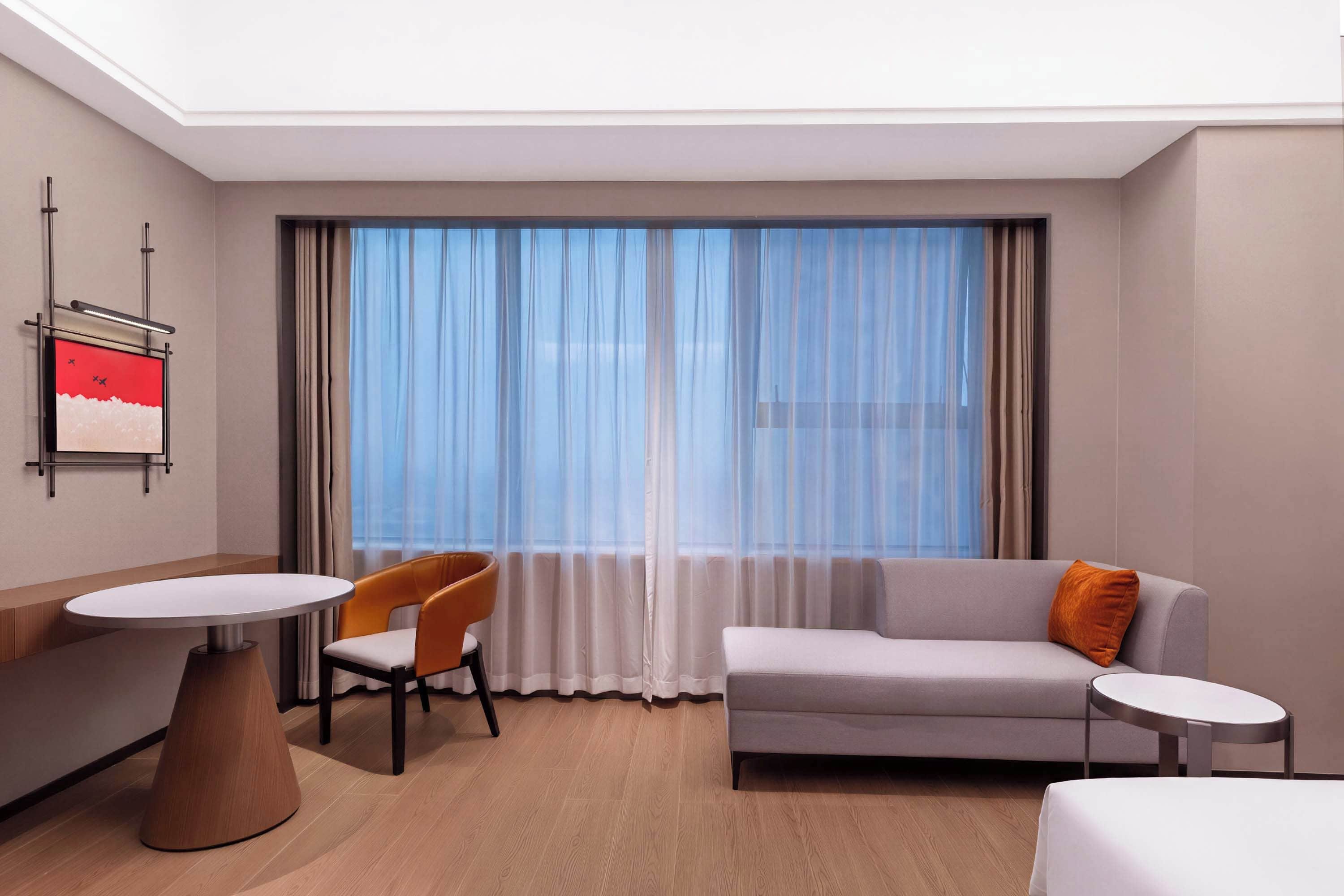 Quarto de hotel com sofá, cadeira, mesa pequena e uma janela grande com cortinas leves.