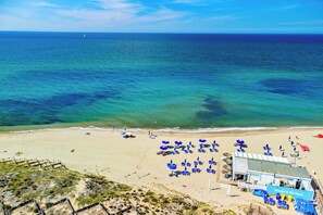 Uma praia com águas azul-claras, areal arenoso e um edifício com a placa 'Welcome to the Beach'.
