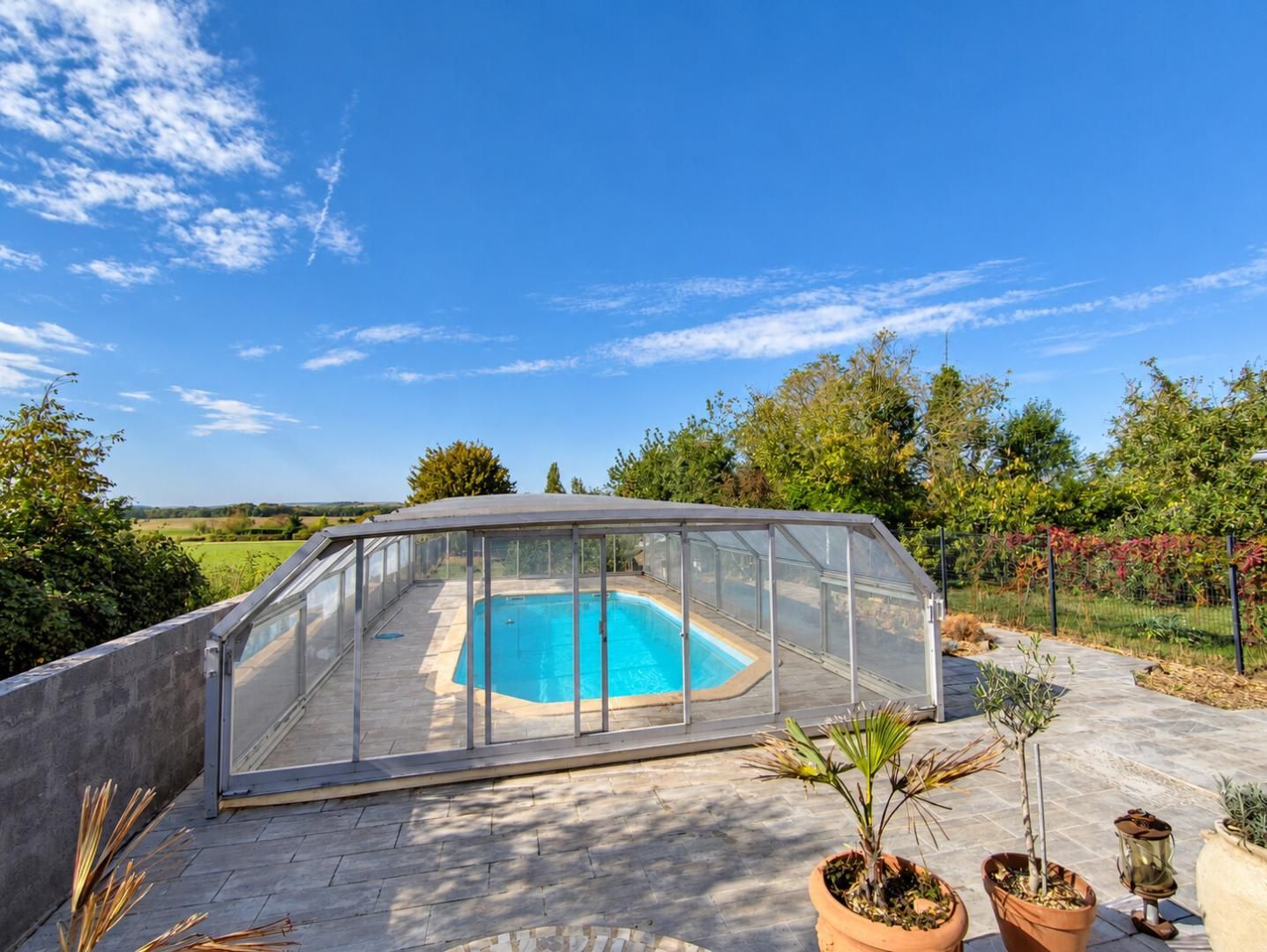 Una piscina con cerramiento de vidrio, rodeada de plantas en macetas y un cielo despejado.