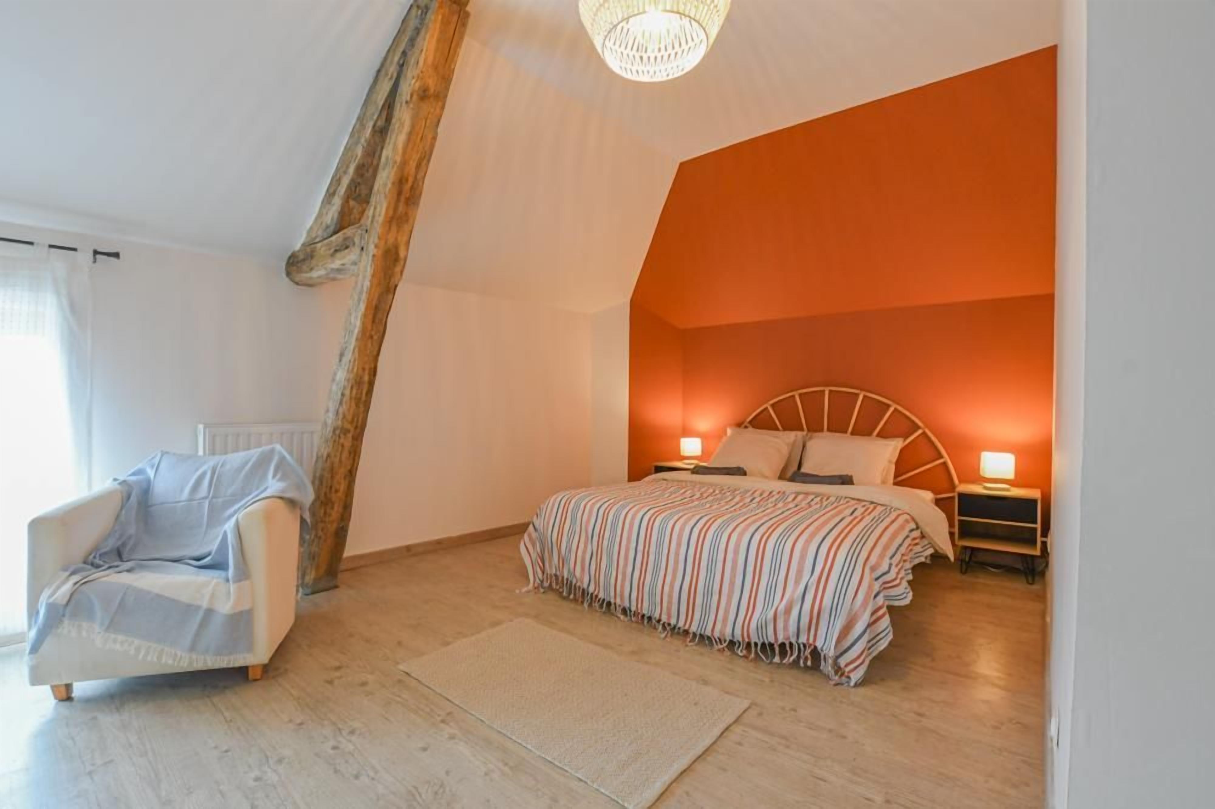 Ein Schlafzimmer mit geneigter Decke, einem Bett mit gestreifter Bettdecke, einem Nachttisch mit Lampe und einem Sessel mit Decke.