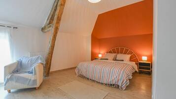 Ein Schlafzimmer mit geneigter Decke, einem Bett mit gestreifter Bettdecke, einem Nachttisch mit Lampe und einem Sessel mit Decke.