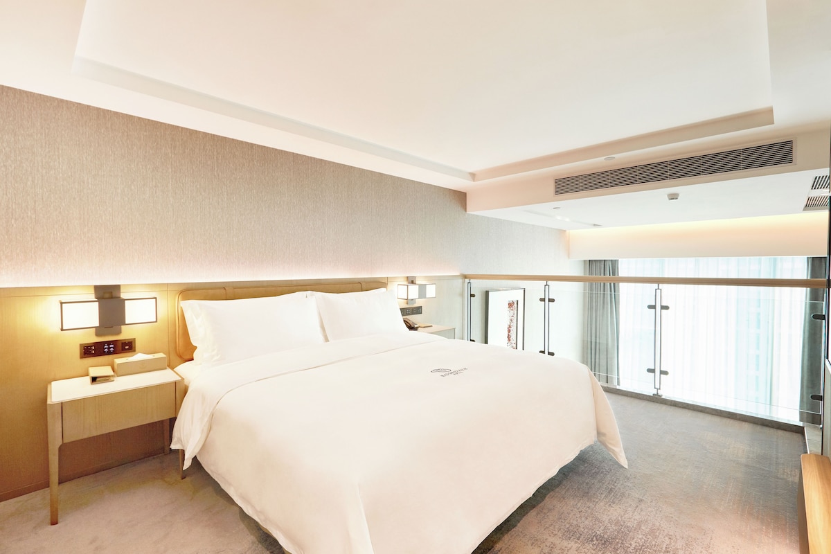 A hotel room with a bed, bedside tables, a nightstand, a window with curtains, and a ceiling vent.
