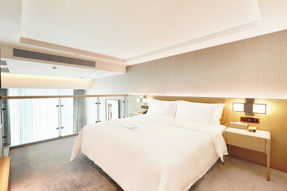 A hotel room with a large bed, bedside tables, a nightstand, a window with curtains, and a view of the outside.