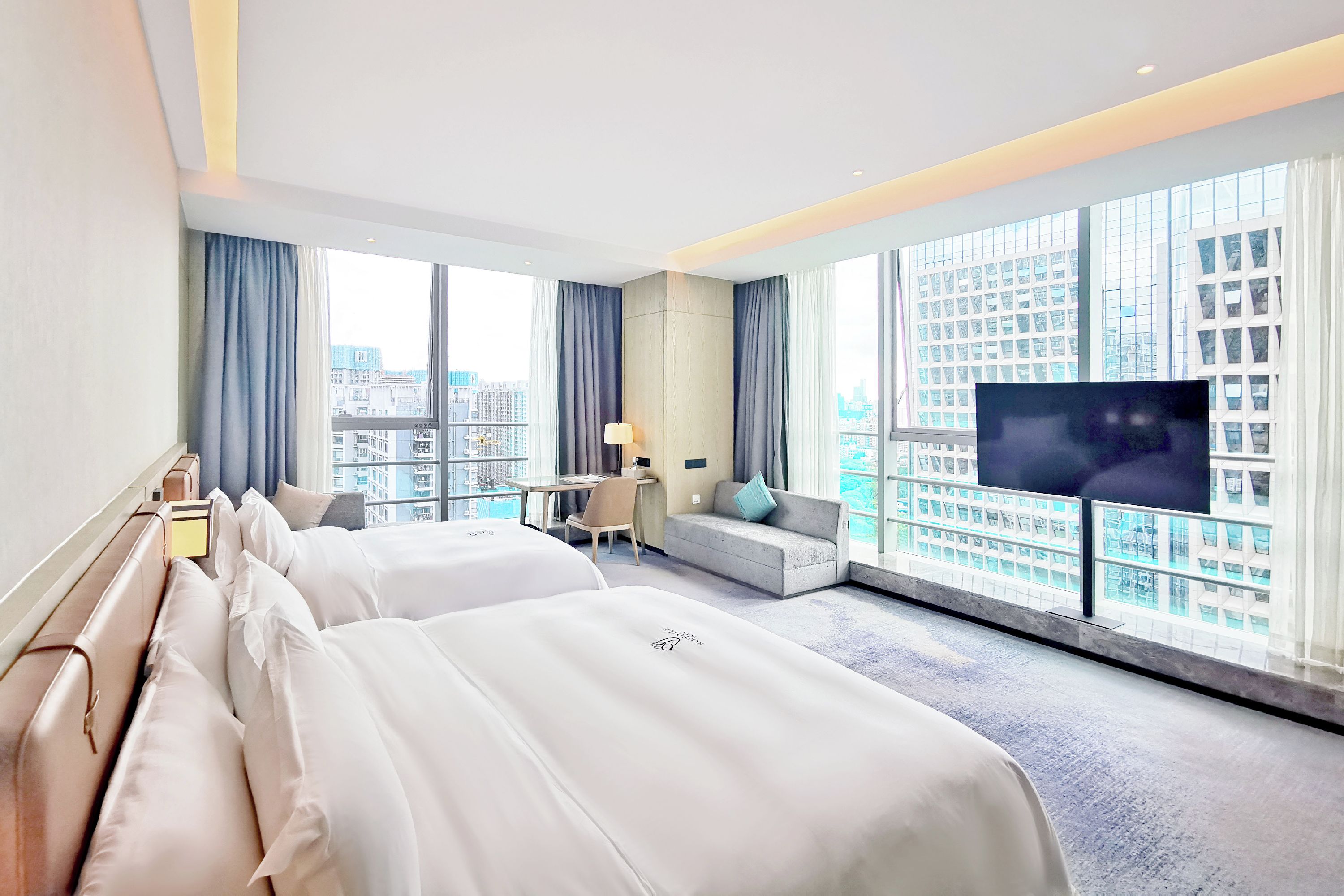 Habitación de hotel con dos camas, un escritorio, una silla, un sofá, un televisor y ventanas grandes con vistas a la ciudad.