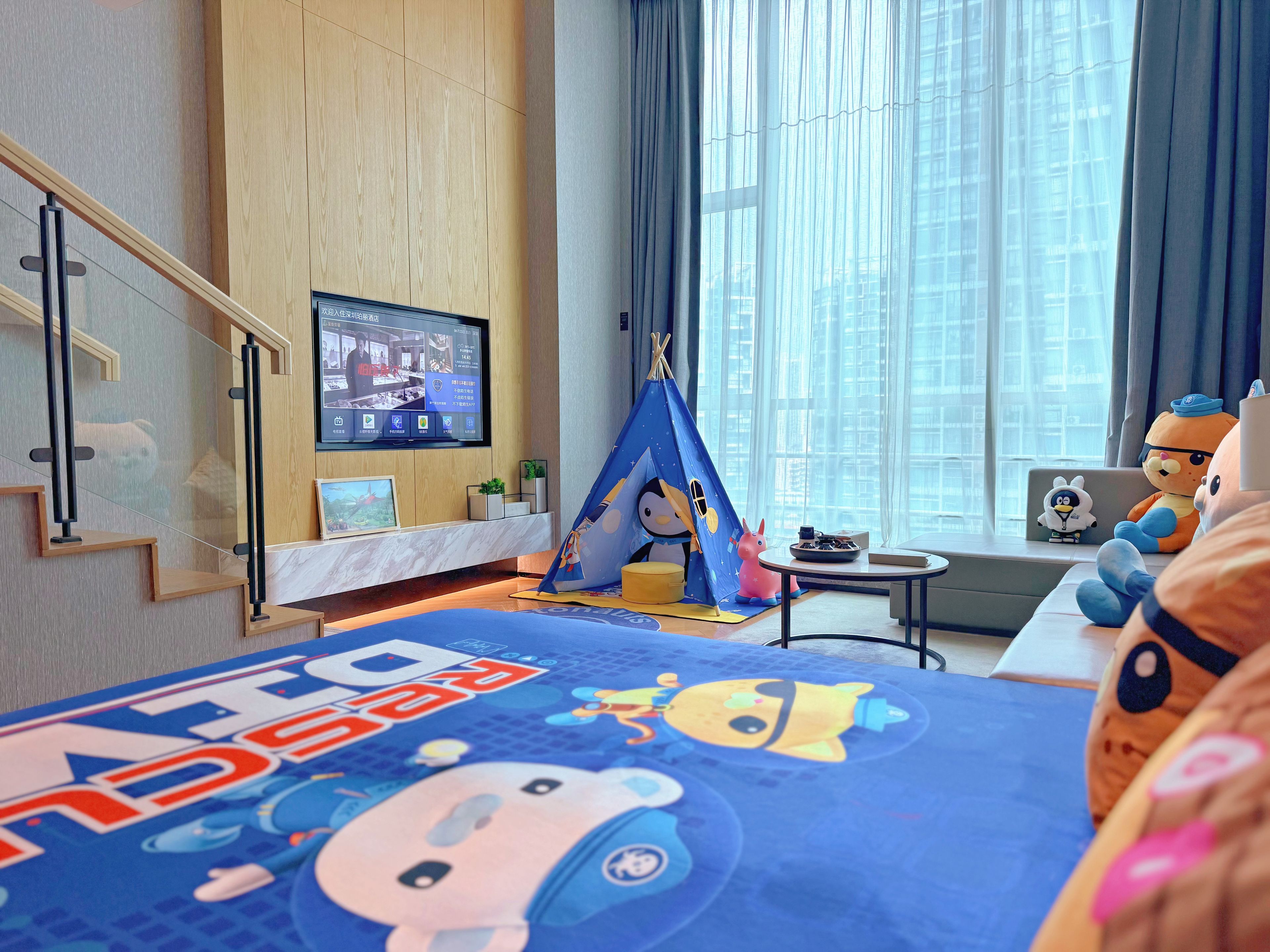 Una habitación de hotel moderna con una cama grande, un televisor, una ventana con cortinas, una mesita pequeña y una zona de juegos con forma de tienda de campaña.