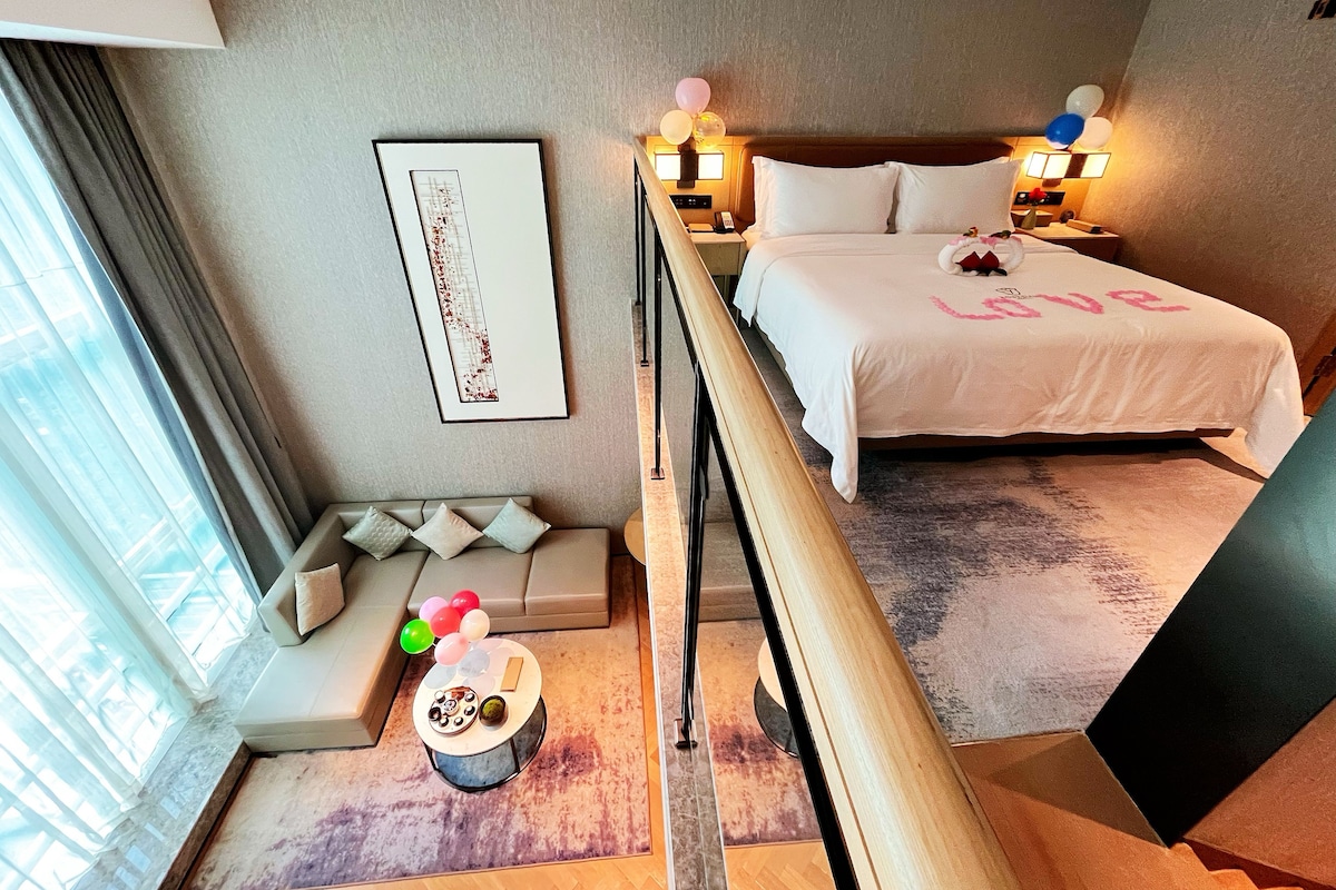 A hotel room with a bed, bedside table, lamp, framed artwork, and a view of the city.