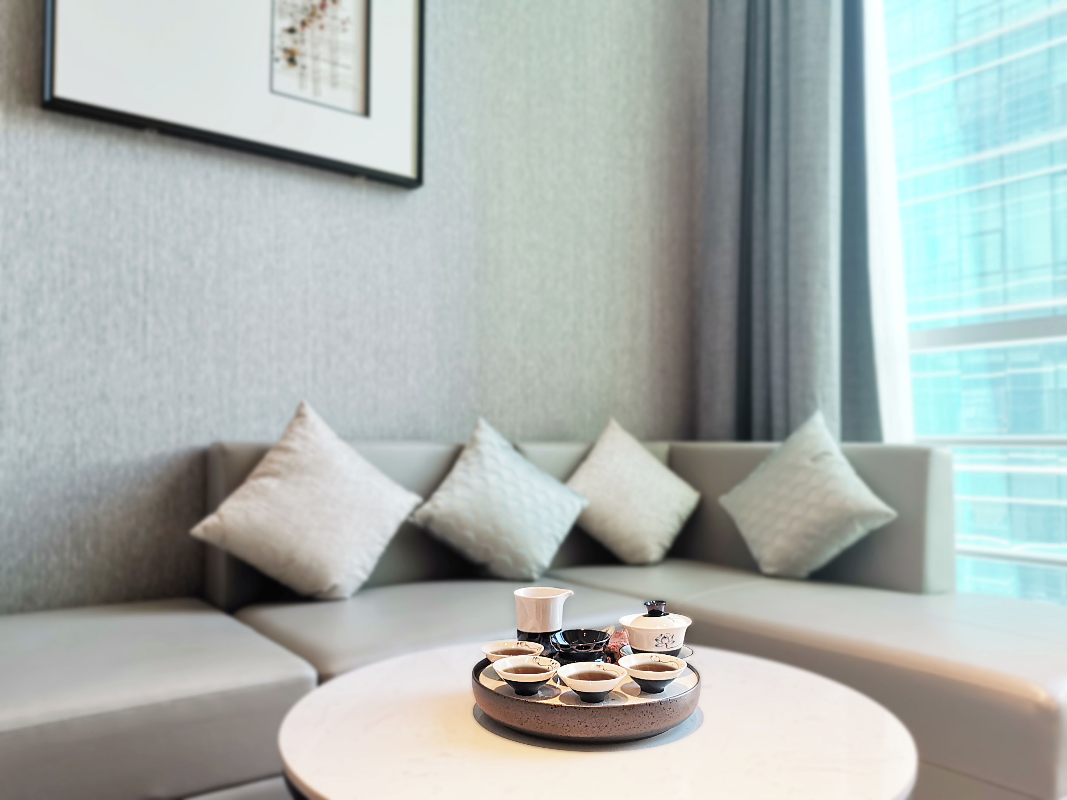 Una sala moderna con una mesa redonda que muestra una tetera y una colección de tazas, un sofá gris con cojines, y un cuadro en la pared.