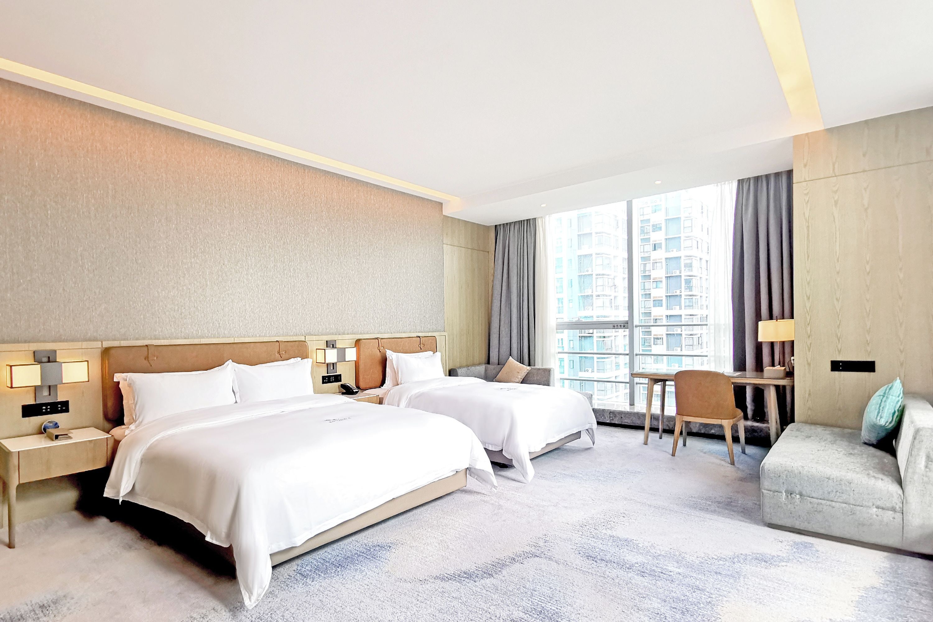 A hotel room with two beds, a desk, a chair, and a view of a building through the window.