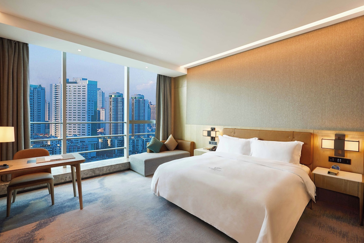 A hotel room with a large bed, a desk, a chair, and a view of a cityscape.