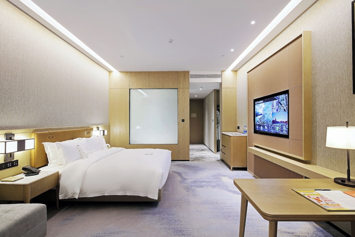 A hotel room with a bed, bedside table, lamp, TV, and a desk with a chair.