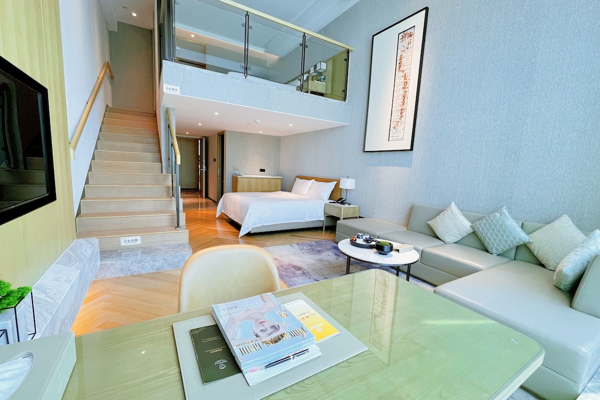A modern hotel room with a bed, a sofa, a dining table, and a staircase leading to an upper level.