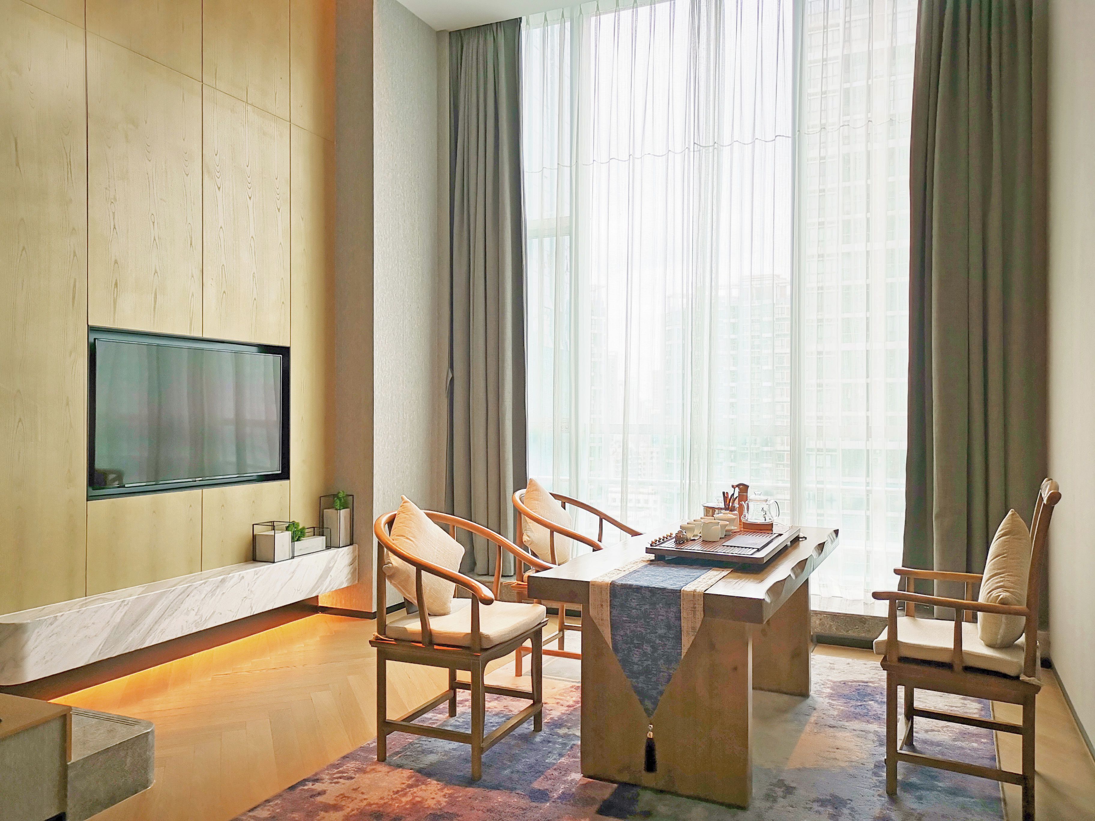 A modern living room with a flat-screen TV mounted on a wooden wall, a dining table with chairs, and a large window with sheer curtains.