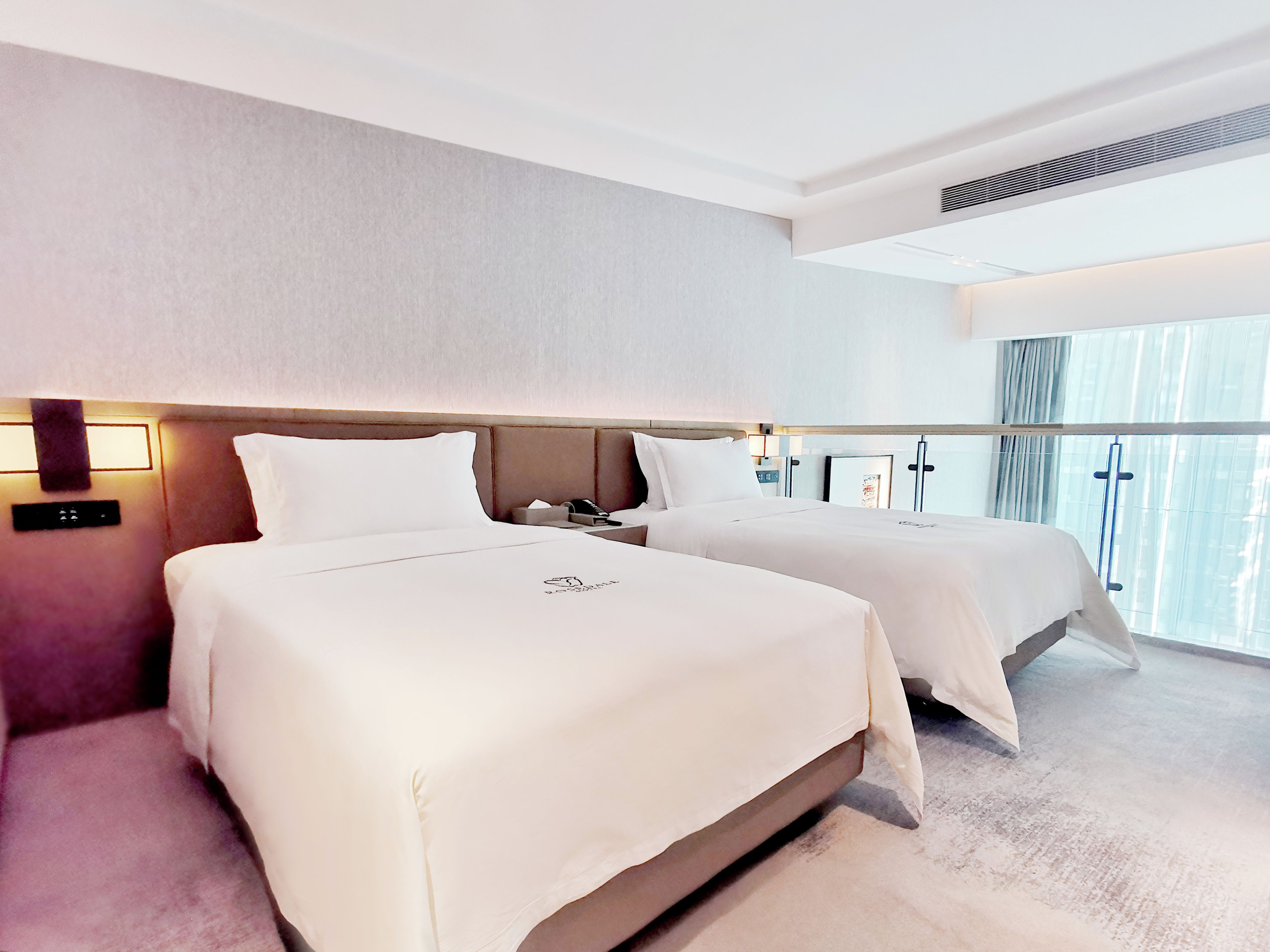 A hotel room with two beds, a nightstand, a wall-mounted lamp, and a large window with curtains.