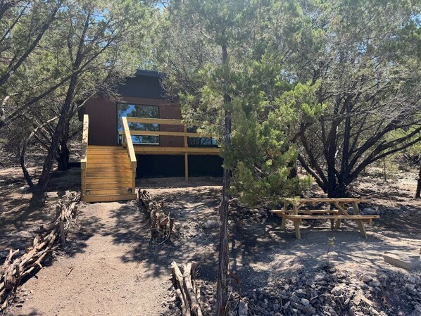 Una cabaña rústica con terraza de madera, rodeada de árboles y un camino de tierra.