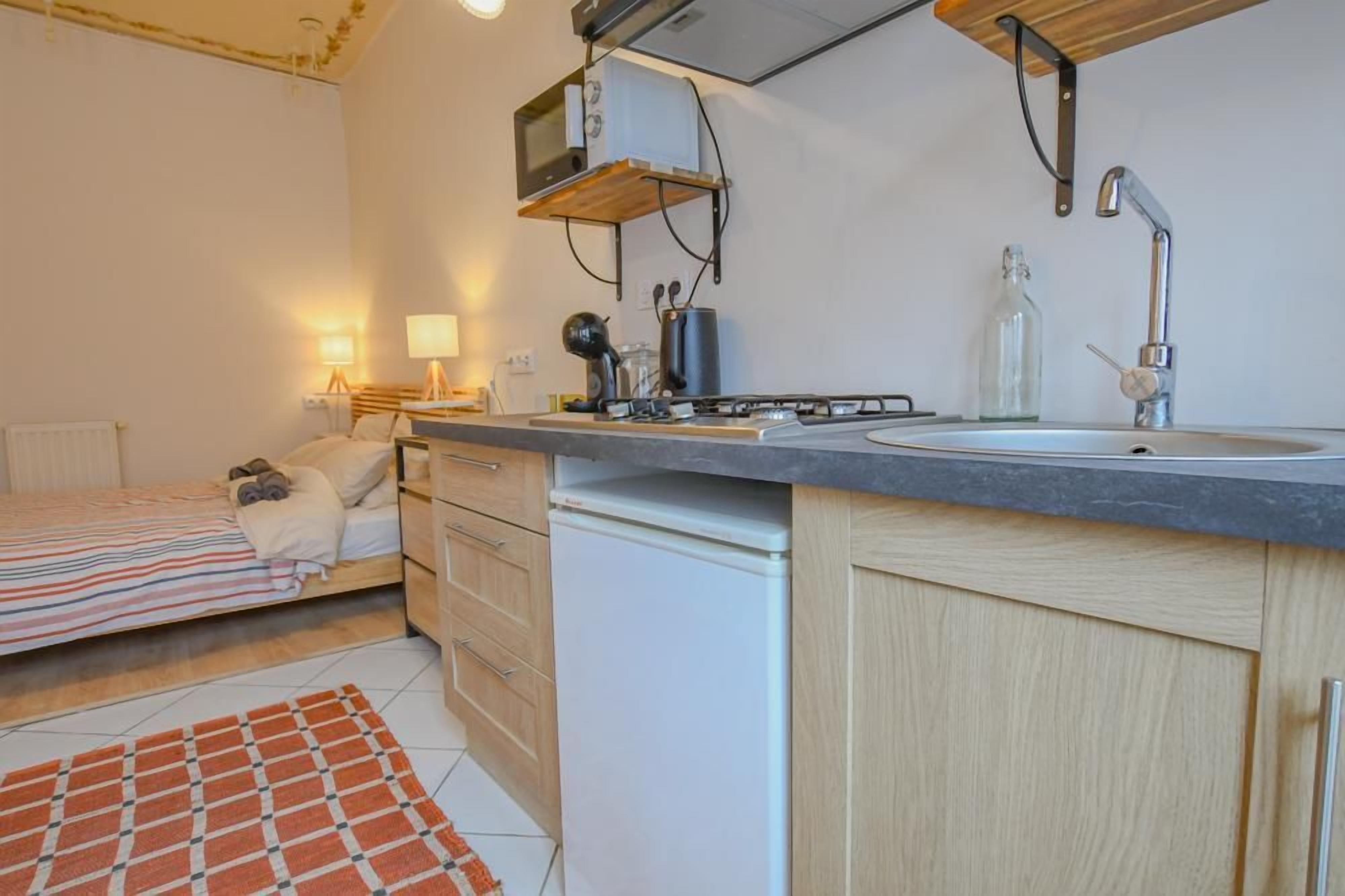 A compact kitchen with a stove, sink, and microwave, a small refrigerator, and a bed with a striped blanket.