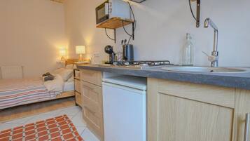 A compact kitchen with a stove, sink, and microwave, a small refrigerator, and a bed with a striped blanket.