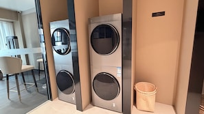 A modern laundry room with two wall-mounted washing machines, a small trash bin, and a seating area with a chair.