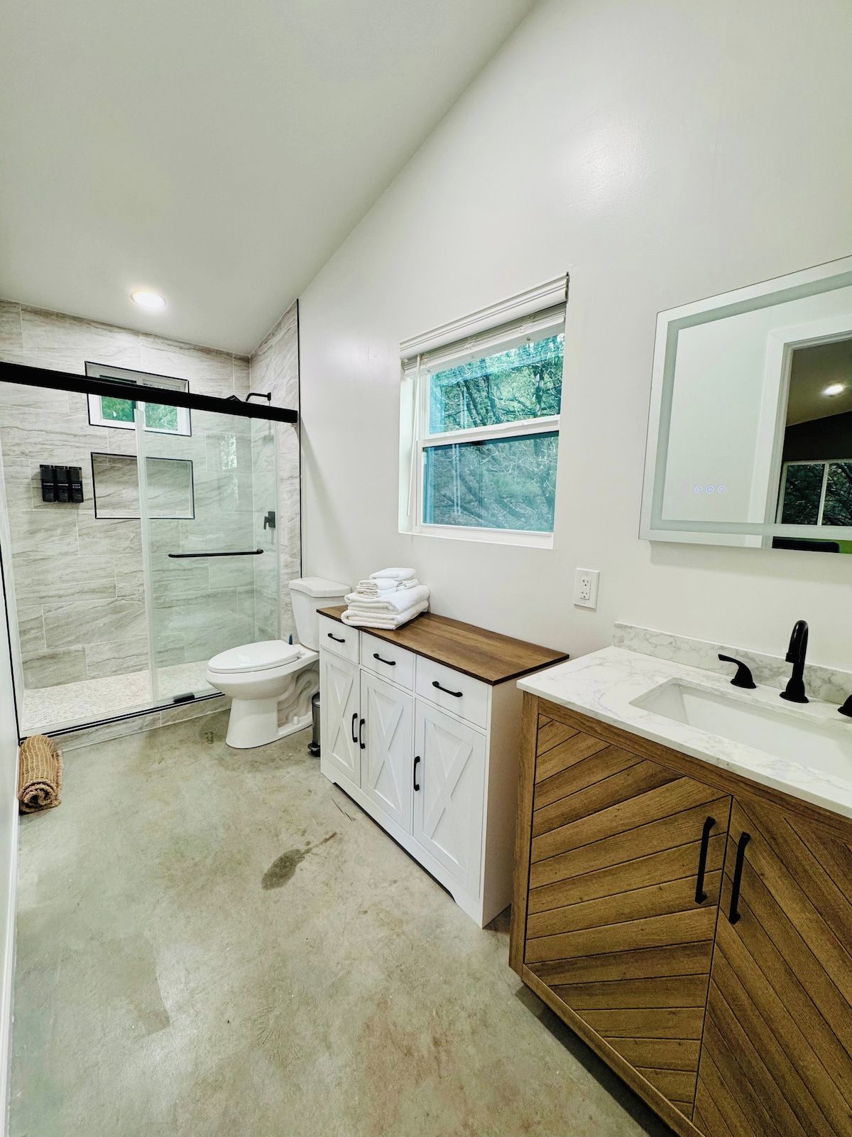 A modern bathroom with a glass shower enclosure, a toilet, a vanity with a sink, and a window.