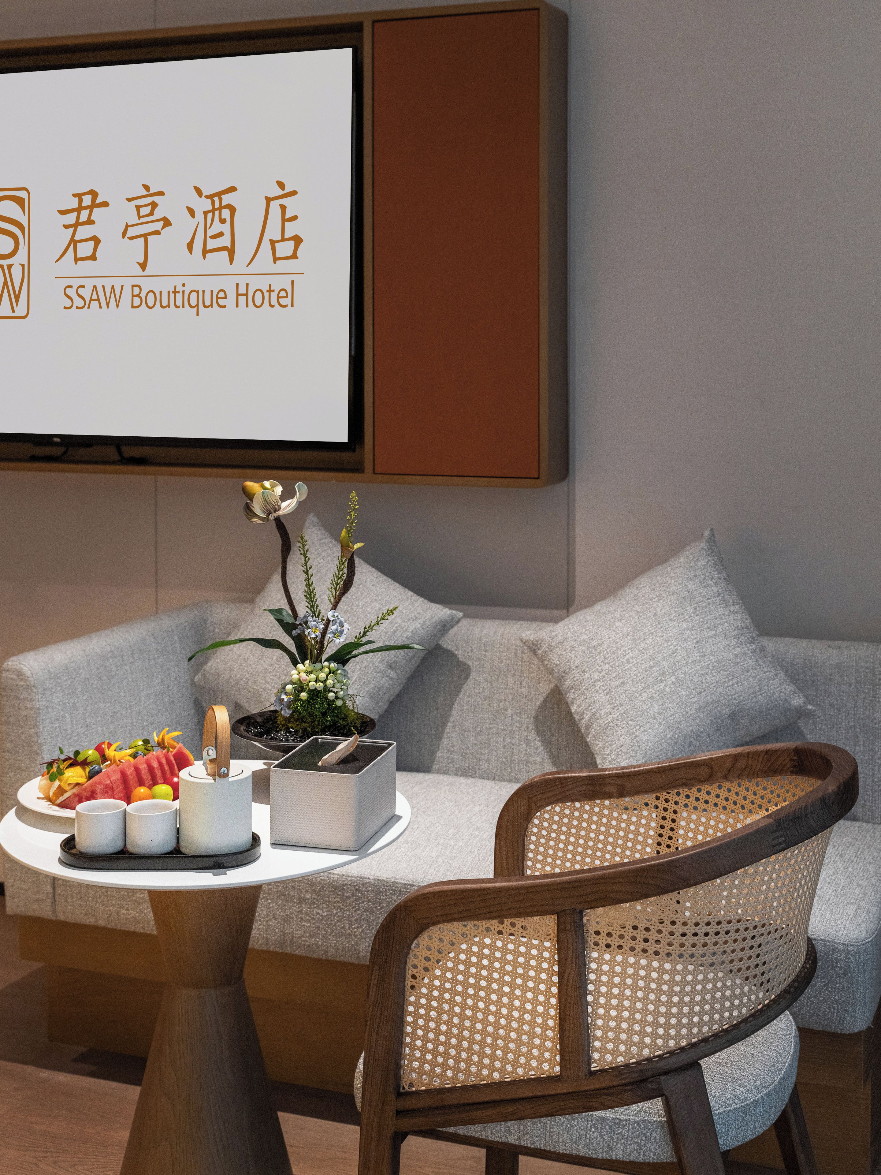 A hotel room with a sofa, a small table with a fruit platter, a potted plant, and a projector screen displaying hotel name.