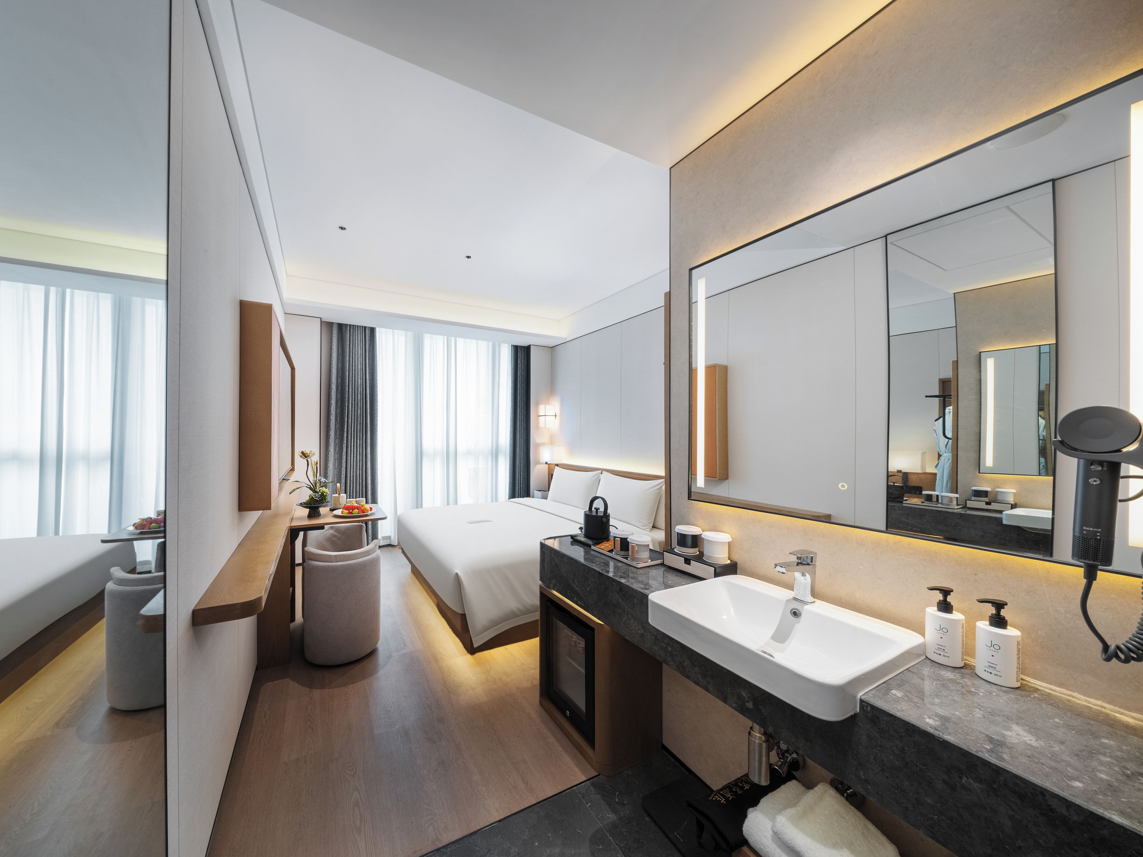 A modern hotel room with a large bed, a sitting area with a table and chairs, a large bathroom with a sink and mirror, and a view of the city through large windows.