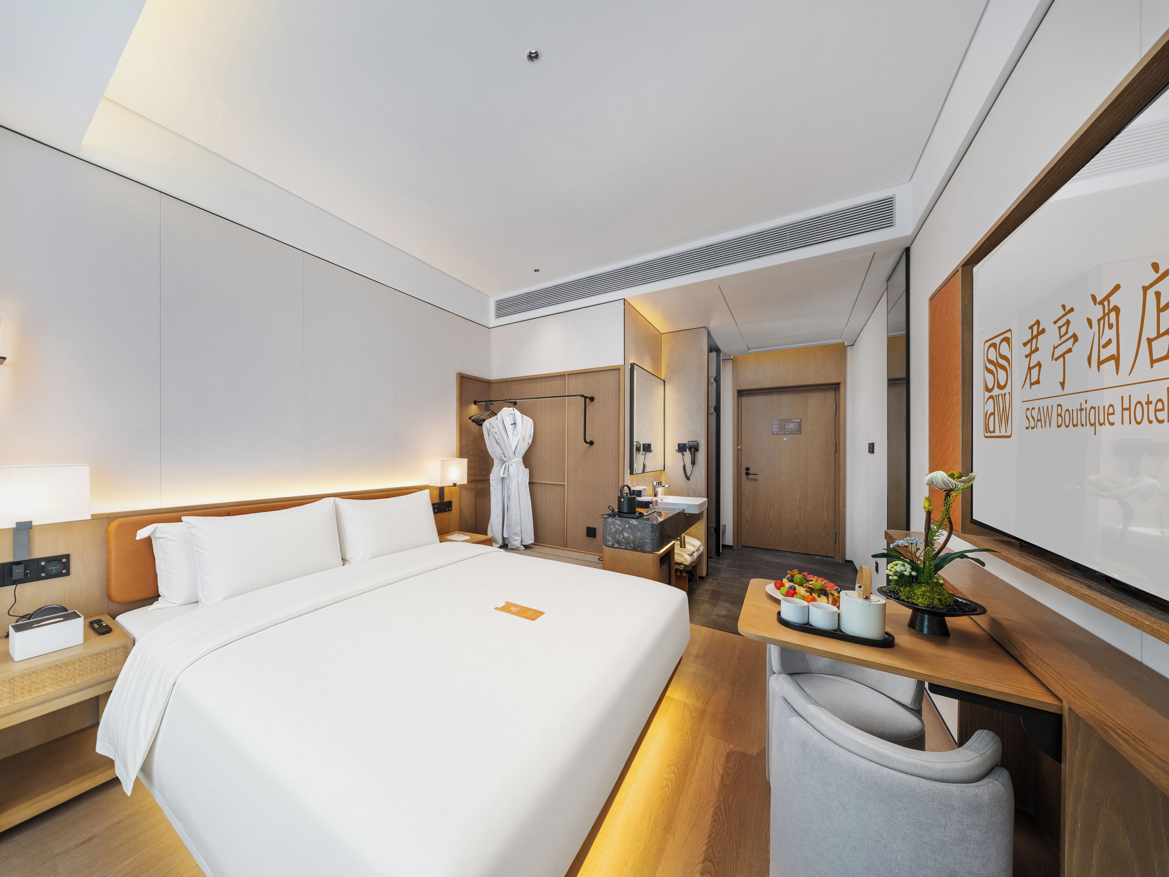 Ein Hotelzimmer mit einem großen Bett, einem Nachttisch mit Lampe, einem Kleiderhaken mit Bademantel, einer kleinen Sitzecke mit Sessel und einem Empfangstresen mit Schild.