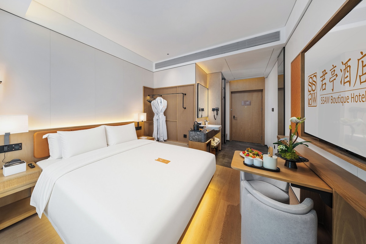 A hotel room with a large bed, a bedside table with a lamp, a robe hanging on a rack, a small seating area with a chair, and a reception desk with a sign.