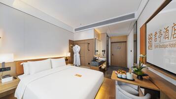 Ein Hotelzimmer mit einem großen Bett, einem Nachttisch mit Lampe, einem Kleiderhaken mit Bademantel, einer kleinen Sitzecke mit Sessel und einem Empfangstresen mit Schild.