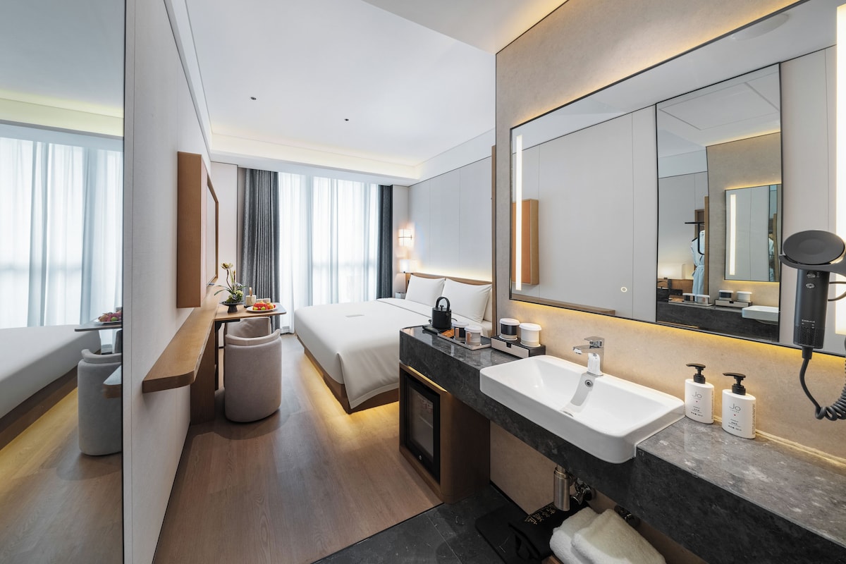 Een moderne hotelkamer met een groot bed, een zithoek met een tafel en stoelen, een ruime badkamer met een wastafel en spiegel, en uitzicht op de stad door grote ramen.