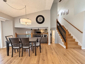 A spacious dining area with a high ceiling, a staircase with wooden railings, and a modern kitchen in the background. - Immaculate Modern Oasis - Huge Backyard (Mead)