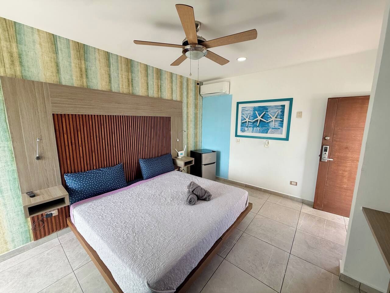 Une chambre d’hôtel avec un lit, un ventilateur de plafond, une table de chevet, un bureau et une porte.