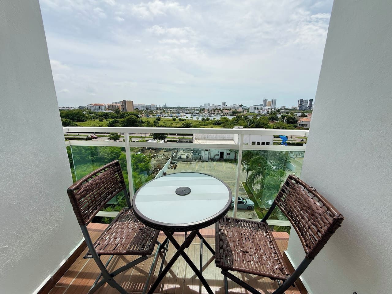 Un balcone con un tavolo e due sedie, affaccia su un panorama urbano con edifici e spazi verdi.