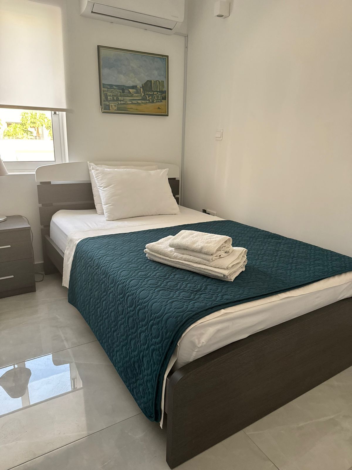 Une chambre à coucher avec un lit, une table de chevet, une fenêtre et un tableau accroché au mur.