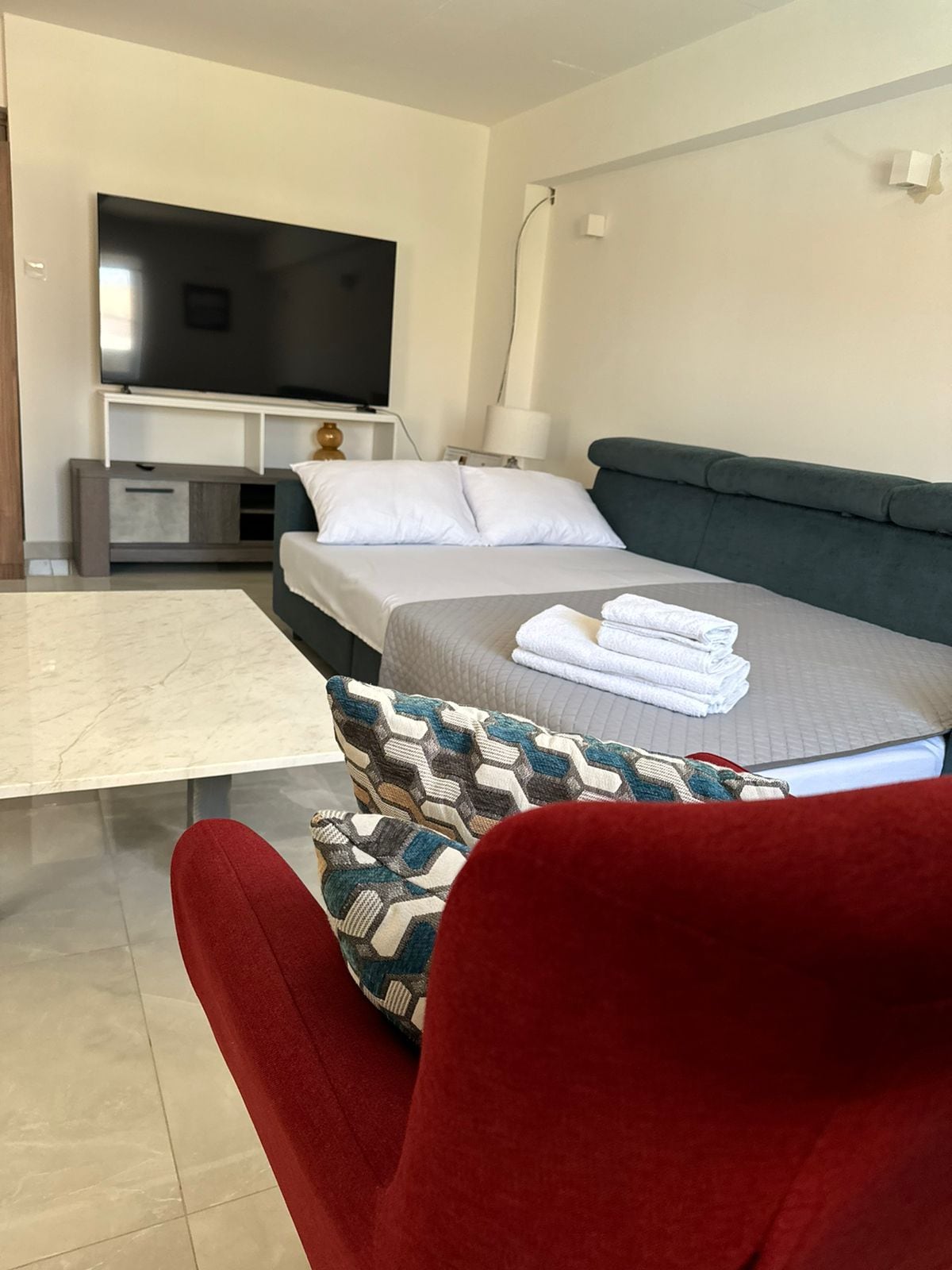 A modern hotel room with a bed, a TV, a sofa, and a red chair.