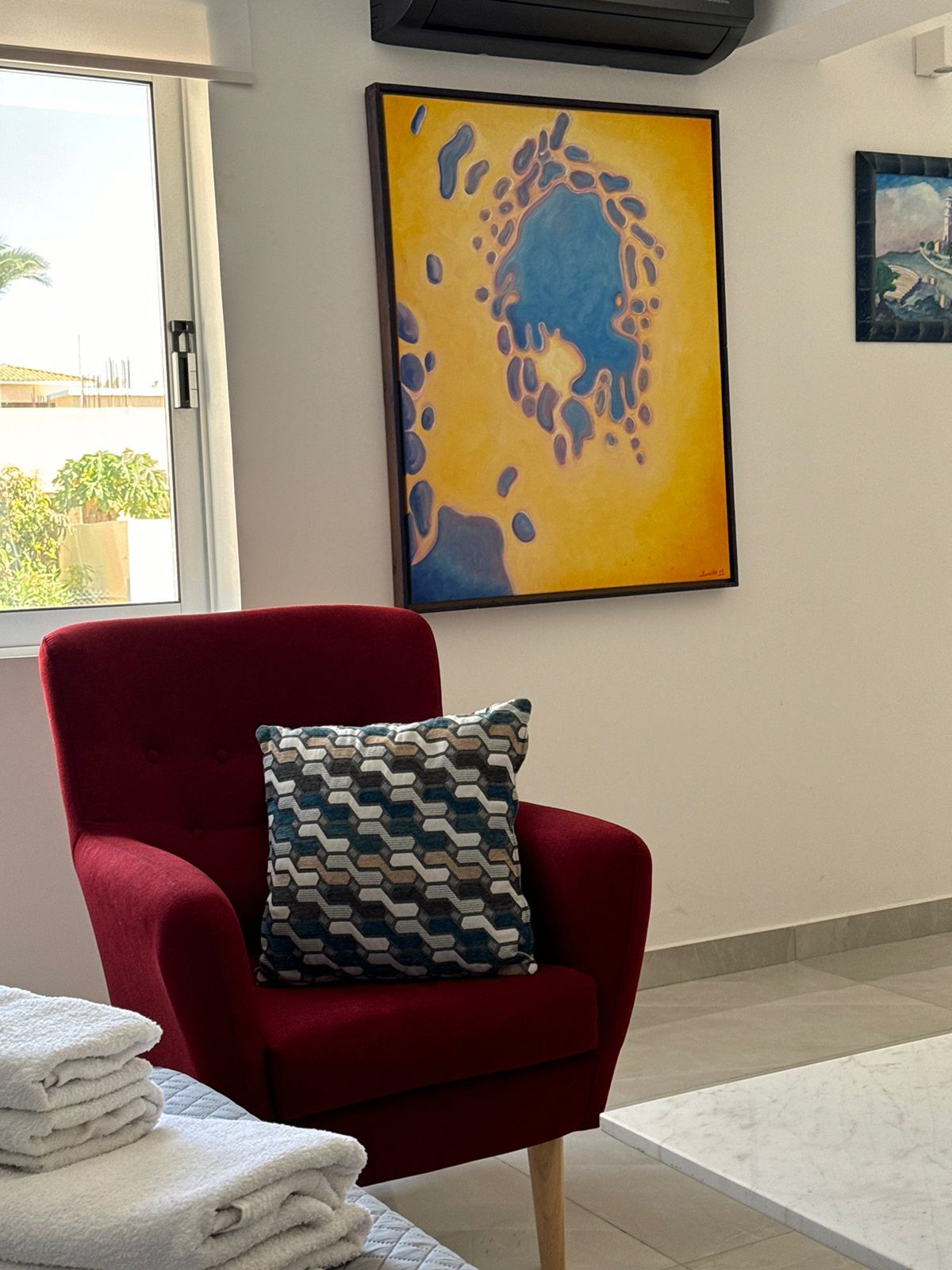 Un fauteuil rouge agrémenté d’un coussin à motifs, une pile de serviettes blanches et un tableau accroché au mur.