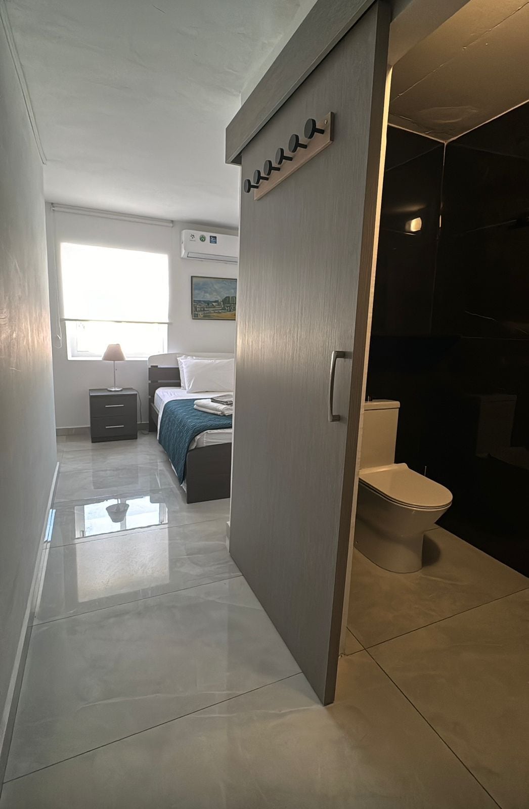 A modern hotel room with a bed, a bedside table, a lamp, a window, and a bathroom with a toilet and sink.