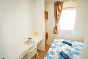 Habitació petita i ordenada amb un llit individual, una taula de treball i una finestra amb cortines.