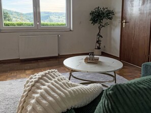 Ein Zimmer mit einem runden Tisch, einem Heizkörper, einer Topfpflanze und Blick auf Grünfläche.