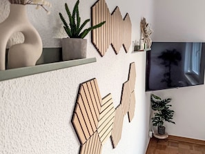 Een moderne woonkamer met een grote flatscreen televisie aan de muur, decoratieve houten wanddecoratie, een potplant en een klein tafeltje met een vaas.