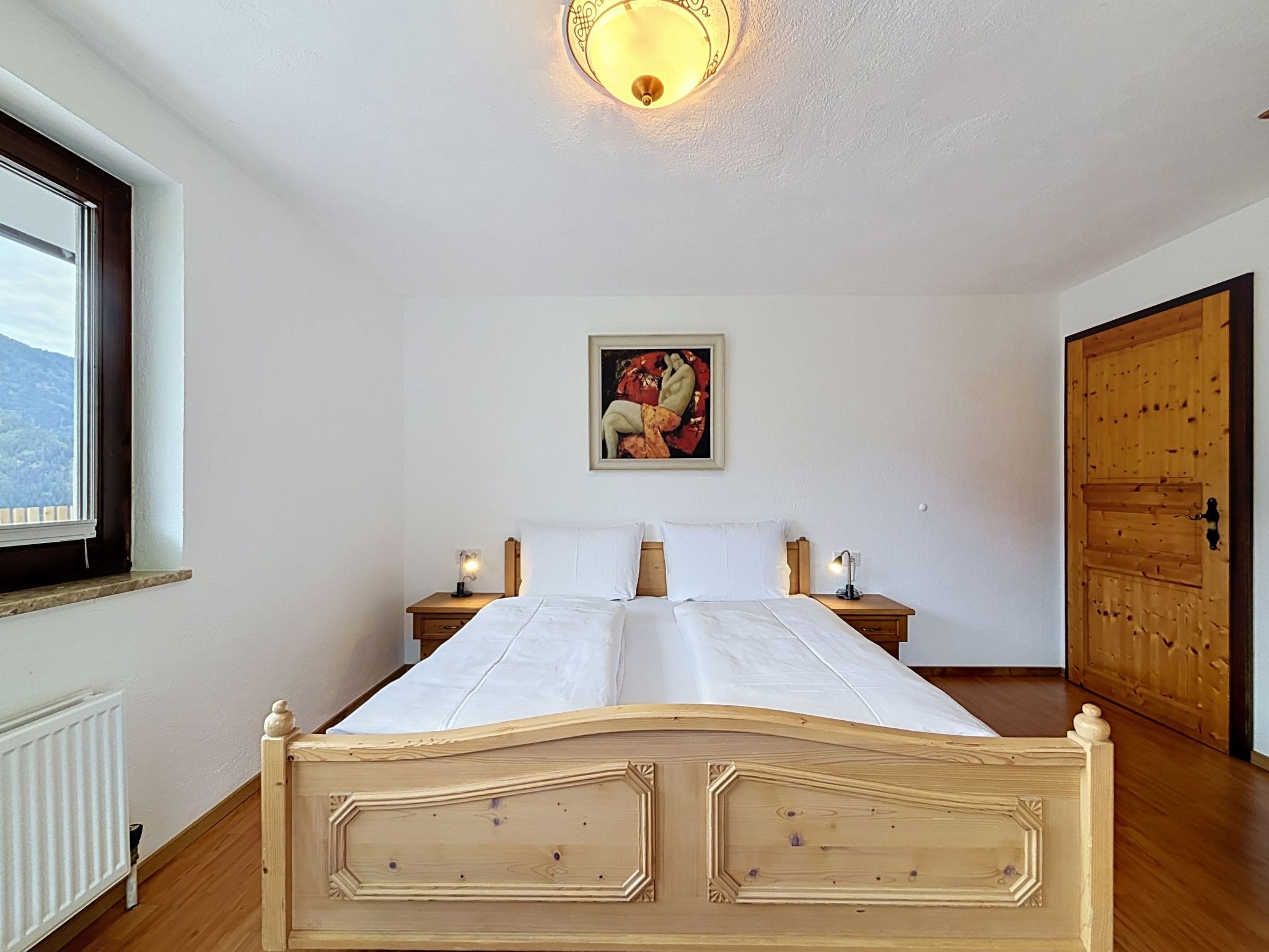 A bedroom with a wooden bed, white linens, a painting on the wall, a window, and a wooden door.