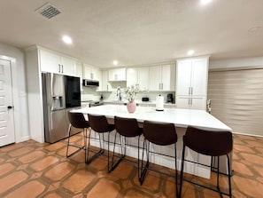 Une cuisine moderne avec des armoires blanches, un réfrigérateur en acier inoxydable, un grand îlot central doté de tabourets et un vase décoratif.