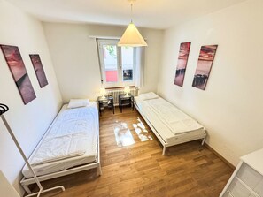 A small, well-lit room with two single beds, a desk with a chair, a radiator, and artwork on the walls. - Apartment  by Interhome (Zurich)