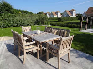 Ein Holz-Essplatz im Freien mit Tisch und Stühlen, eine Glasvase mit Kerze und ein gepflegter Garten mit Hecken und Rasen.