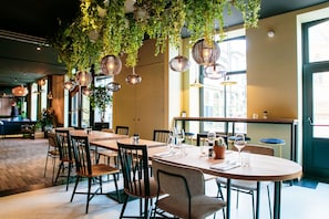 L’intérieur d’un restaurant présente une longue table conviviale, des chaises individuelles, des plantes suspendues et des luminaires suspendus.