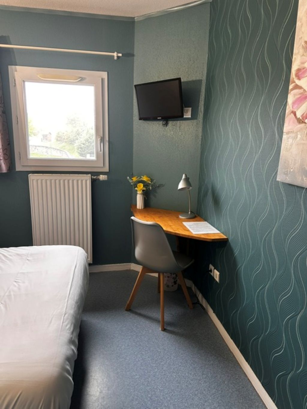 Een slaapkamer met een bed, een bureau met stoel, een radiator, een raam en een televisie aan de muur.