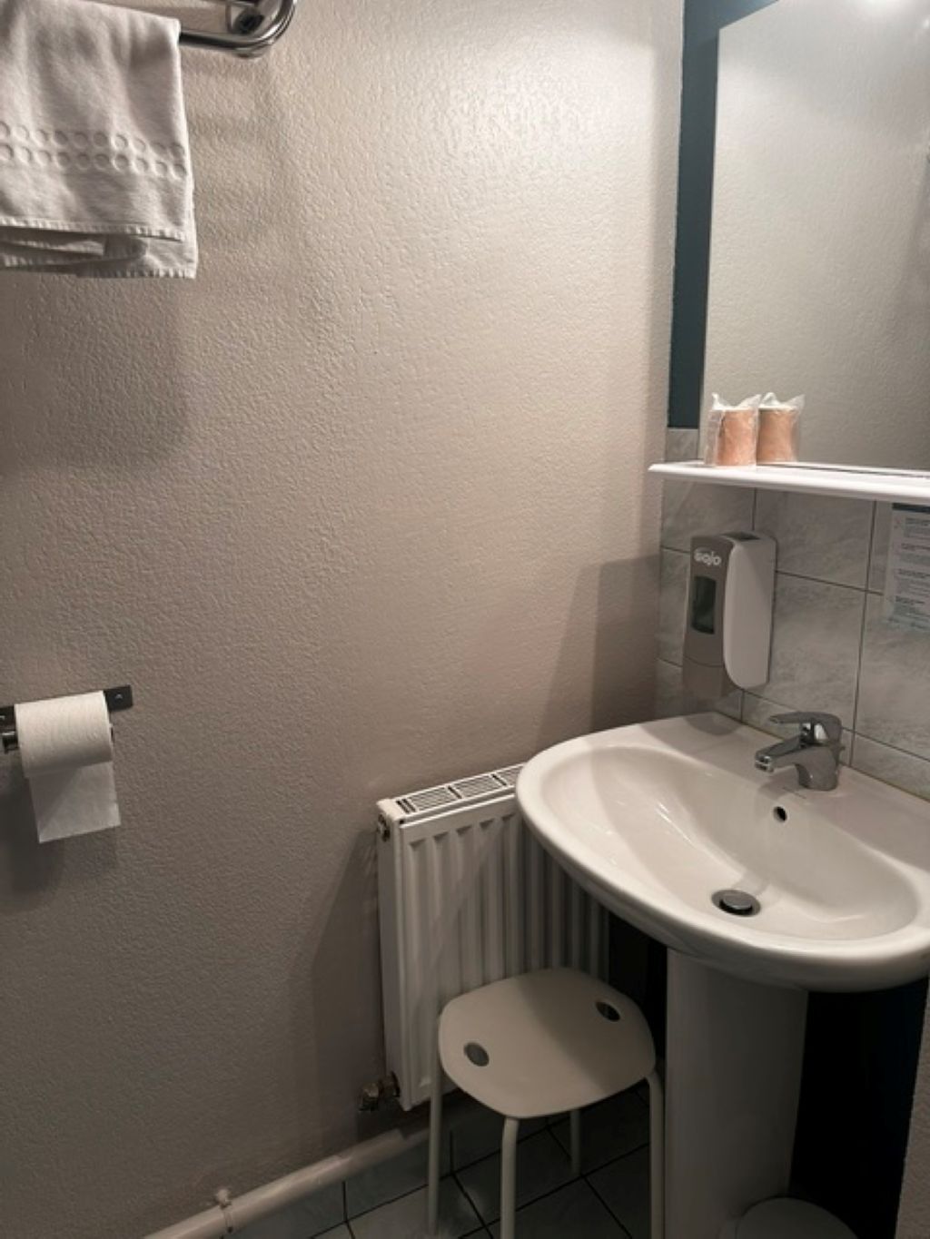 Ein Badezimmer mit Waschbecken, Toilette, Handtuchhalter und einem kleinen Hocker sowie einer Heizung.