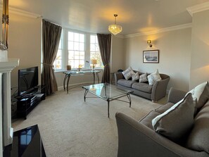 A living room with a sofa, a glass coffee table, a television, and a chandelier. - 2 bedroom accommodation in Whitby (Whitby)