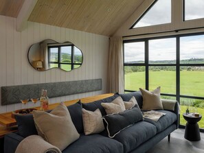A modern living room with a dark blue sofa, a wooden coffee table, and a large window offering a view of a green field. - 2 bedroom accommodation in Ravensworth (Ravensworth)