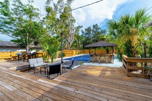 Una terraza de madera con mobiliario exterior, una parrilla y un área de piscina rodeada de árboles.