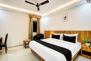 A hotel room with a bed, a desk, a chair, a ceiling fan, a nightstand, and a wall-mounted picture. - Townhouse GRAND ADDRESS HOTEL  (Ghaziabad)