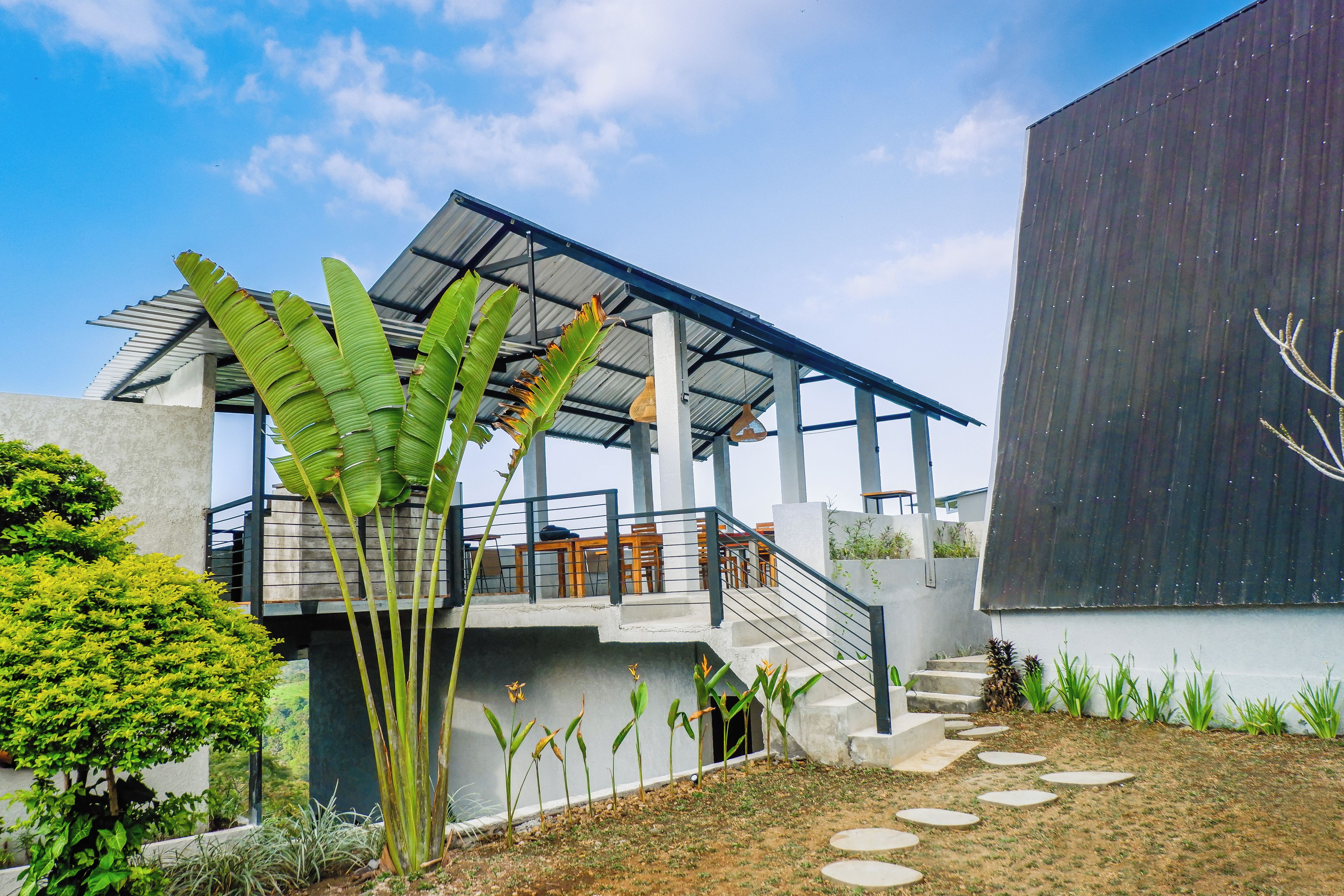 A modern building with a covered outdoor seating area, a staircase, and a garden with various plants.